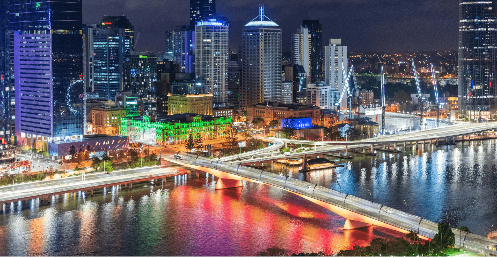City skyline at night with illuminated buildings, river, and bridge.