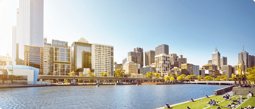 A serene river scene with individuals relaxing on the grass, framed by a bustling city in the background.
