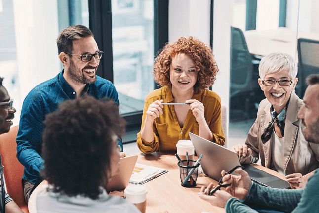 Diverse group of coworkers smiling and discussing ideas during a business meeting around a circular wooden table in a bright, modern office space.