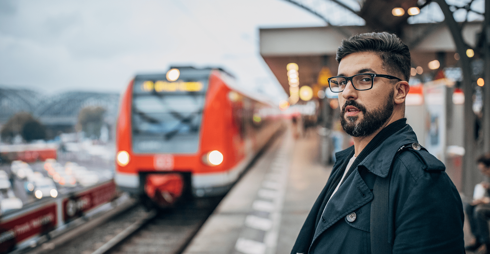 A man with glasses and a beard stands in front of a train, looking towards the camera with a neutral expression.