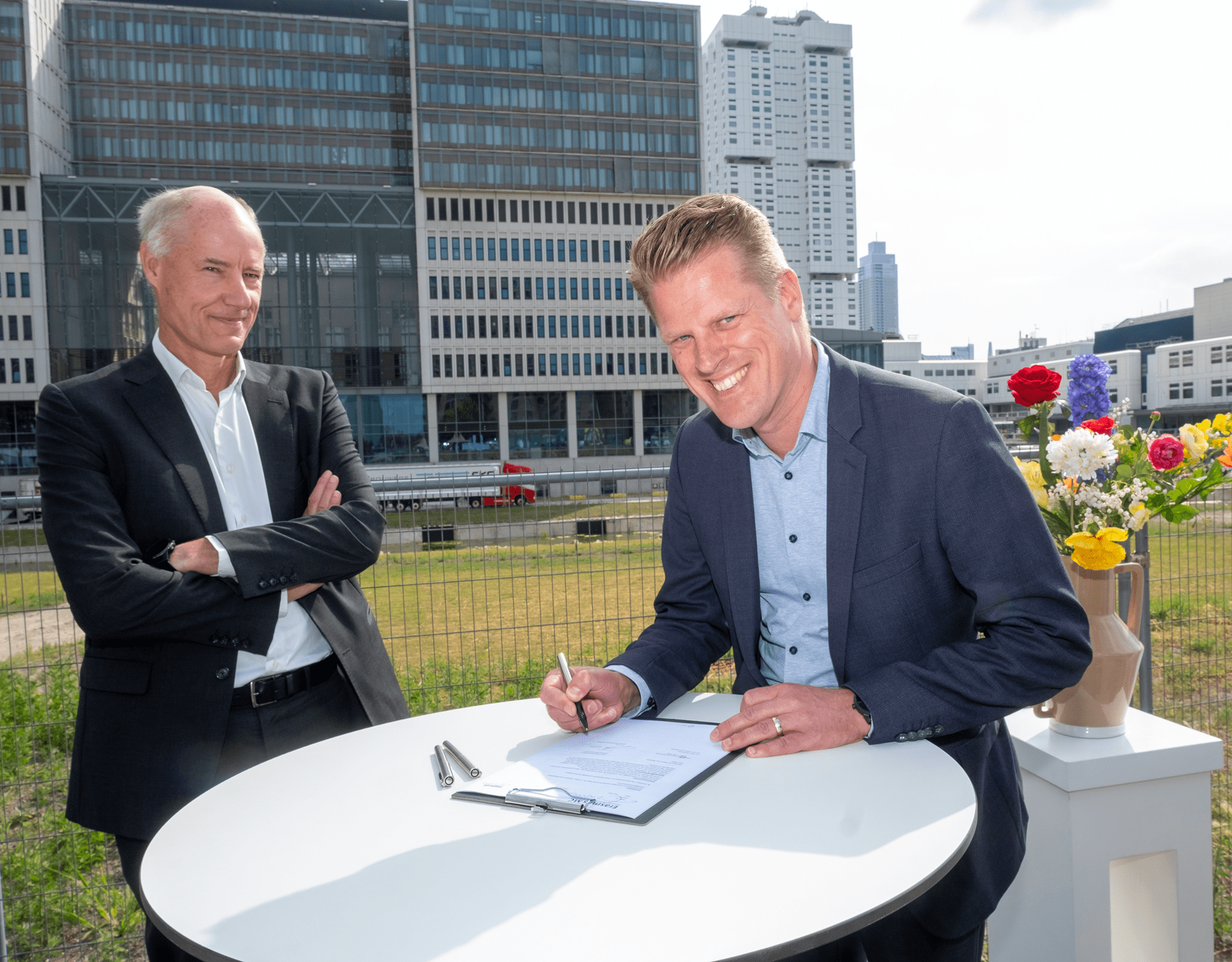Two men signing documents in front of a modern building, engaged in a formal agreement process.
