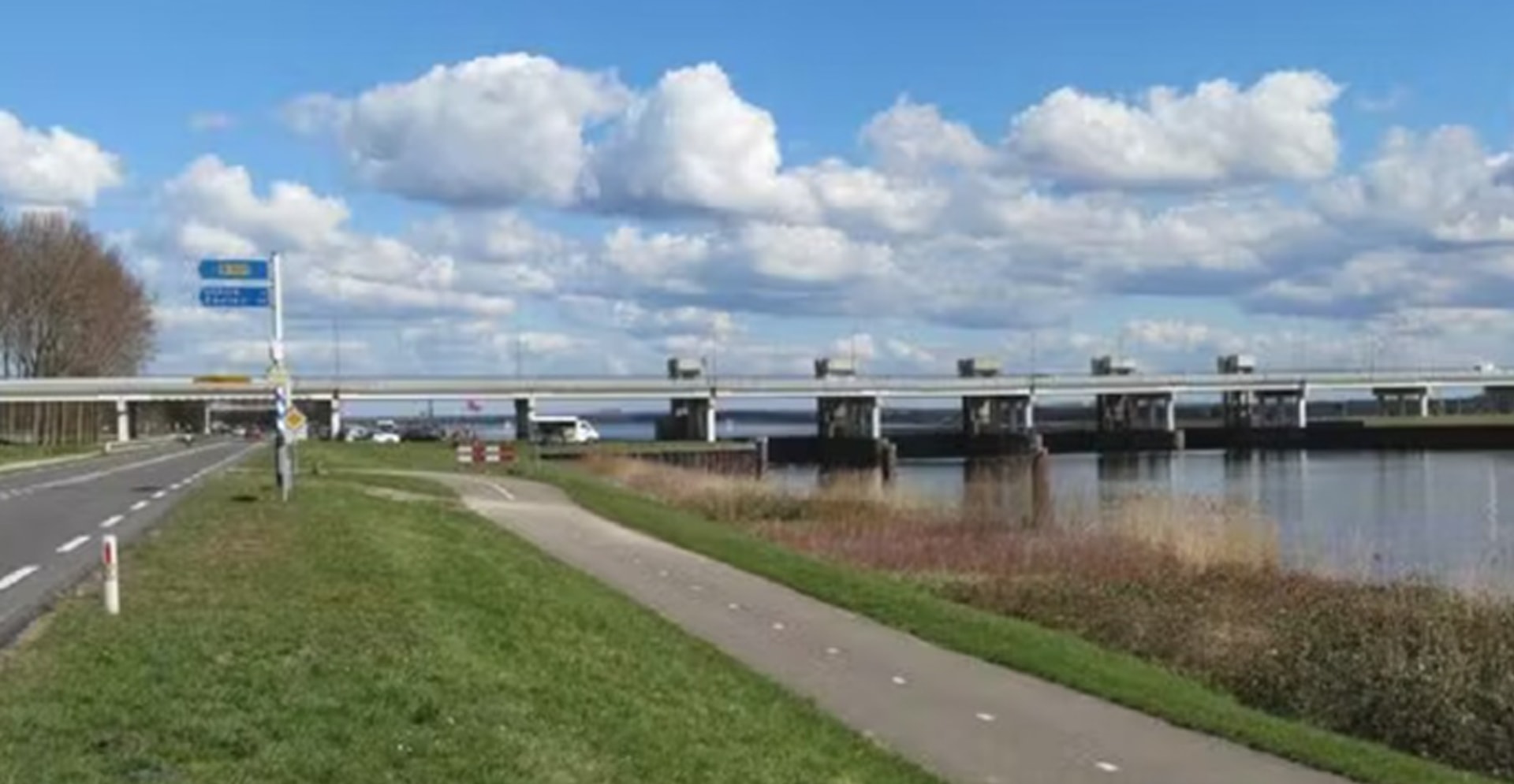 A bridge crosses a river, with a road visible on the far side, framed by trees and a bright sky.