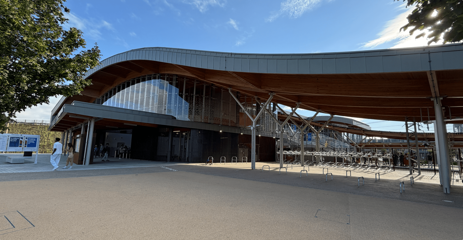 Modern train station with a curved, wood-accented roof and glass facade. Sunlight casts shadows, creating a serene, spacious atmosphere.