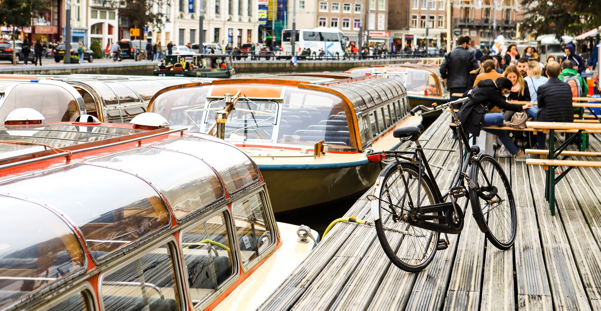 Canal landscape in Amsterdam with boats, a bicycle parked on a wooden on the dock with people on the background.