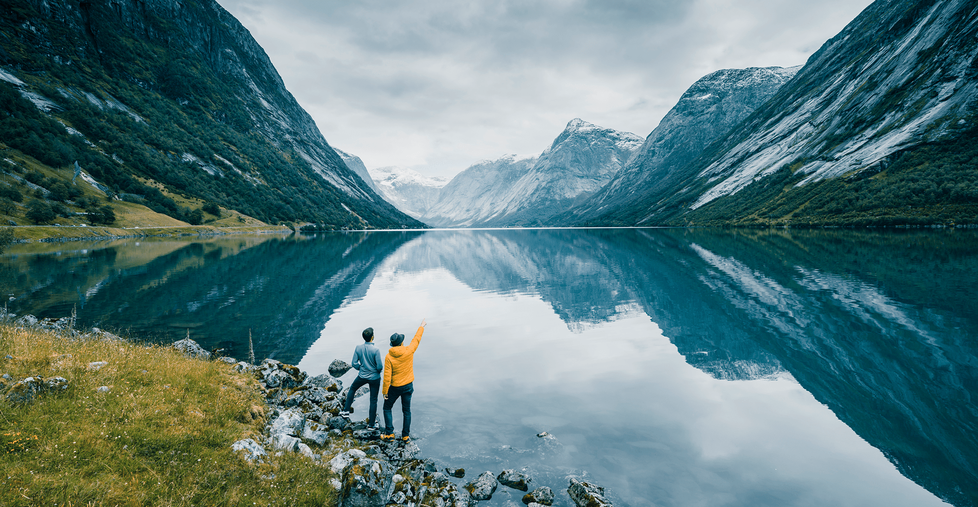 Two people stand on a lakeshore, surrounded by mountains, enjoying the serene landscape.
