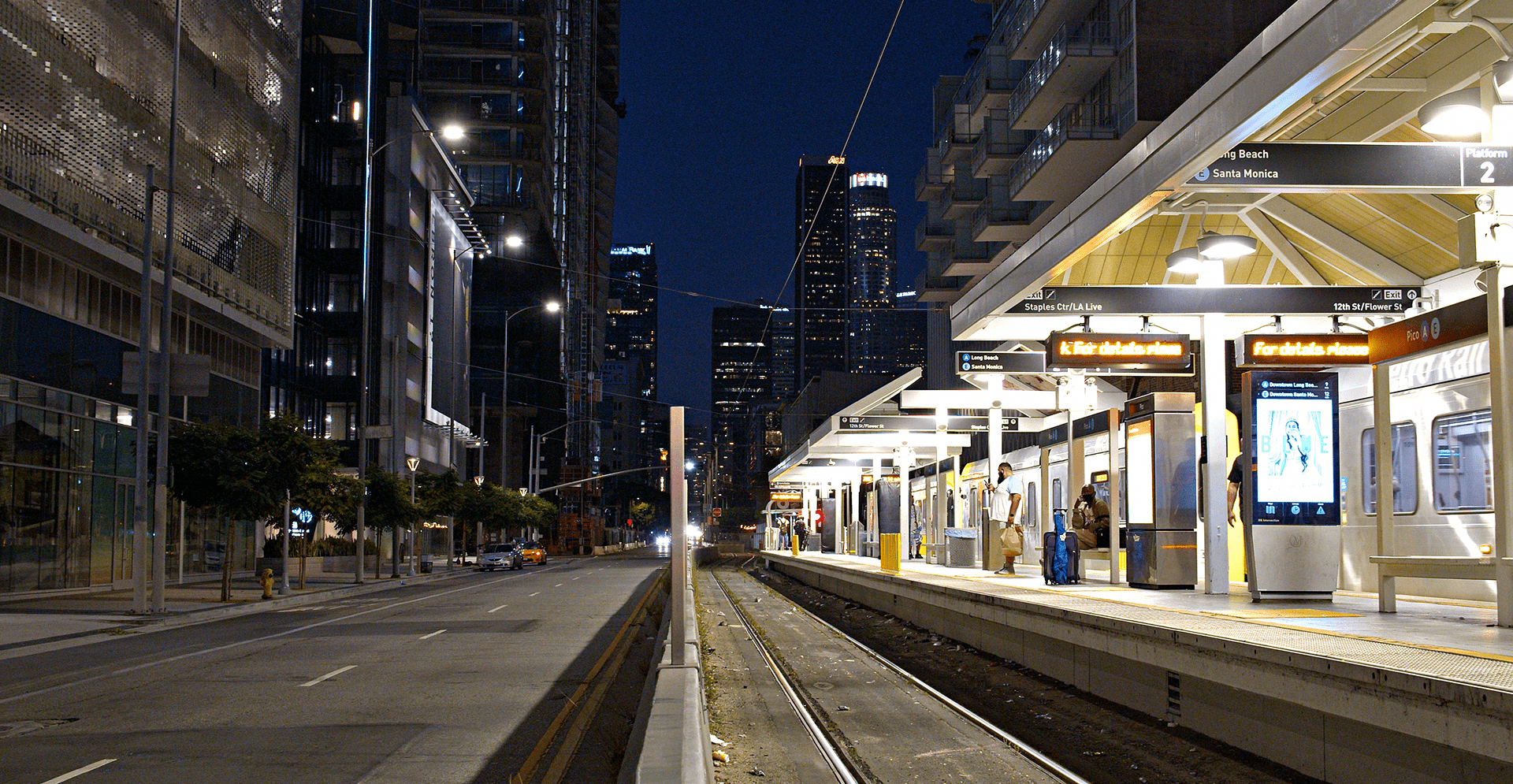 LA Metro station at night with downtown skyline and passengers waiting by train platform.