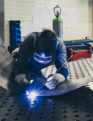 A factory scene showing a welder using a welding torch on steel, surrounded by industrial equipment and safety gear.