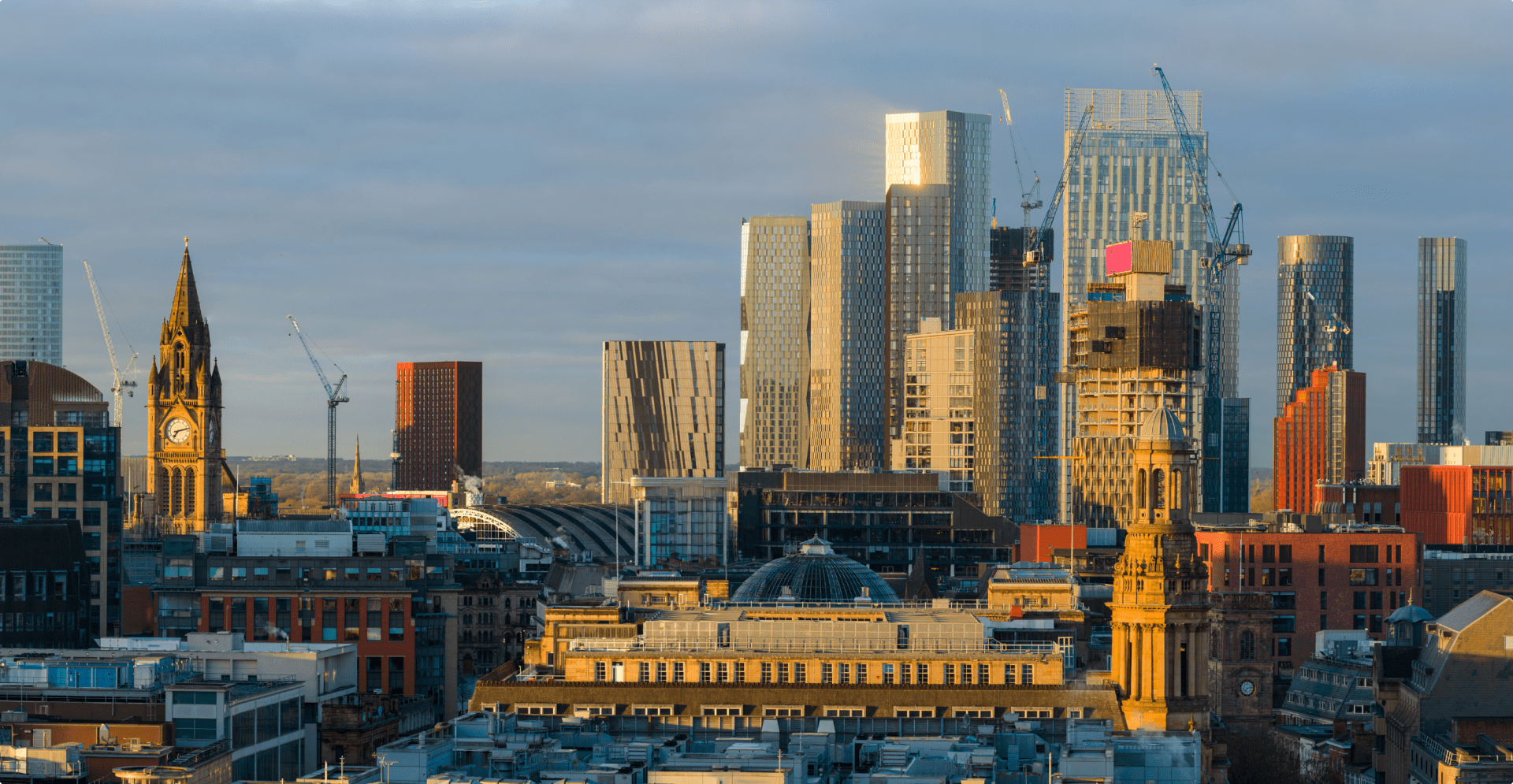 Skyline of a city at golden hour, featuring a mix of historic and modern skyscrapers. A clock tower is prominent, with cranes in the background.