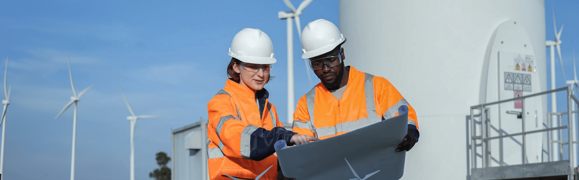 Two workers in safety vests and hard hats stand next to wind turbines, emphasizing safety in a clean energy environment.