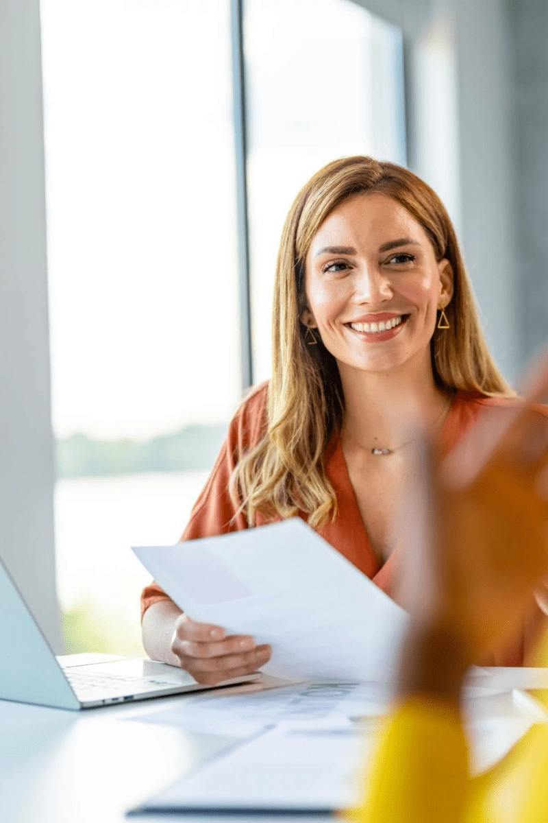 A woman with a bright smile sits at a table, focused on her laptop, embodying a moment of joy and concentration.