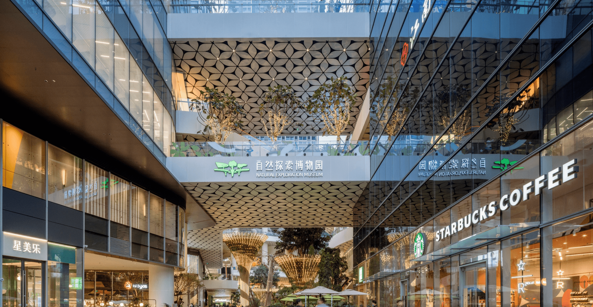 A spacious modern shopping mall interior featuring shops, escalators, and bright lighting with shoppers walking around.