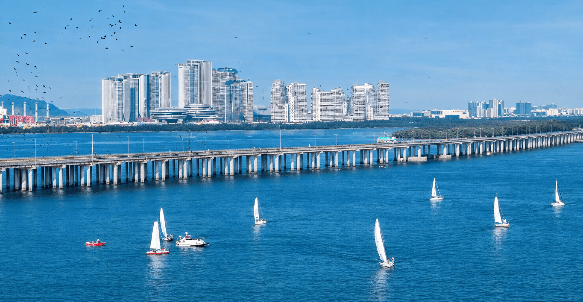 A serene coastal scene with sailboats gliding on blue waters, a long bridge spanning across, and a skyline of modern high-rise buildings under a clear sky.