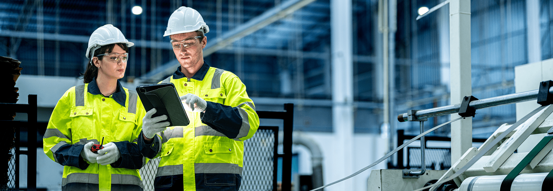 Two safety-equipped workers pose in front of industrial machinery, highlighting safety protocols in a work environment.