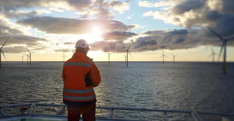 Offshore manual worker standing on helipad with wind-turbines behind him in sunset