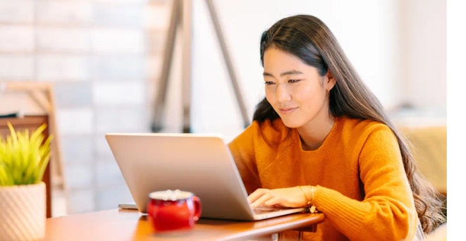 Woman in an orange sweater sits at a table, working on a laptop Woman in an orange sweater sits at a table, working on a laptop